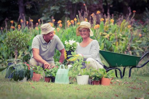 The image depicts a cheerful elderly couple in a well-maintained garden, surrounded by various potted plants, flowers, and greenery. The man is wearing a light-colored hat and a casual short-sleeved shirt, while the woman is also wearing a wide-brimmed hat and glasses, holding a flowering plant. They are seated on the grassy area, with a wheelbarrow and garden tools nearby, indicating active gardening work typical of Mayfair outdoor spaces. The garden features a lush, green lawn with a backdrop of colorful flowering beds, including purple and orange blossoms, and leafy shrubs, creating a vibrant and inviting outdoor environment. The scene is illuminated by natural daylight, suggesting a clear day suitable for outdoor gardening activities, emphasizing landscape maintenance and gardening services offered by Landscapers Mayfair for landscaped gardens and outdoor spaces in the local area.