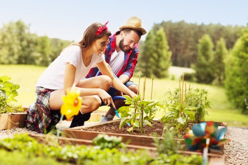 The image depicts a young woman and a man engaged in gardening activities in a well-maintained outdoor garden space. They are kneeling near a raised wooden vegetable bed, which contains small green plants and young seedlings. The woman, wearing casual clothing and a pink headband, is tending to the plants with her hands, while the man, dressed in a plaid shirt and straw hat, observes and assists. The garden bed is edged with gravel, and a decorative bird-shaped garden ornament is visible in the foreground. In the background, there are lush, green trees and an open grassy area under a clear, sunny sky, suggesting a warm weather day. The scene highlights outdoor gardening efforts typical of landscaped residential gardens in the Mayfair area, showcasing fresh greenery and active plant care. Landscapers Mayfair may assist with garden planting, maintenance, and landscaping services to enhance outdoor spaces in the local area, infusing natural beauty and outdoor functionality into private gardens.