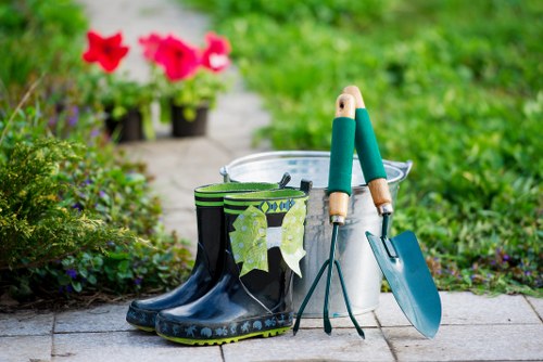 A pair of black rubber gardening boots with green camouflage pattern trim are placed on a paved pathway in a garden, with a pair of garden tools—a trowel and a hand rake—with wooden handles resting against a white plastic bucket nearby. Behind the bucket, there are vibrant pink flowers in pots, and the background features lush green foliage and a well-maintained lawn with a soft, natural lighting suggesting a mild, overcast day. The garden appears to be part of a landscaped outdoor space, potentially in or near Mayfair, with an emphasis on neat, organized plant care and outdoor maintenance as part of professional gardening services provided by Landscapers Mayfair.