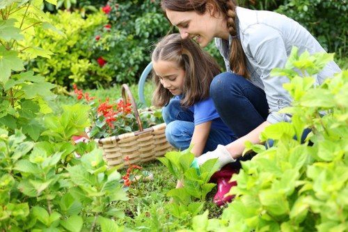 A woman and a young girl are gardening together in a lush, well-maintained backyard. The woman, with long brown hair in a braid, is dressed in a light-colored shirt and dark jeans, crouching down to assist the girl with planting or tending to a flower bed. The girl, wearing a blue top and pink shoes, holds a small garden trowel and is focused on the plants. The garden features vibrant green foliage, including leafy bushes and flowering plants with red blossoms, arranged around a neatly cut lawn with short, dense grass. In the background, a hedge or row of shrubs provides privacy and structure to the outdoor space. The environment appears bright and sunny, suggesting a warm day, with natural light illuminating the scene. This setting exemplifies outdoor gardening activities suitable for residential yards in the Mayfair area, emphasizing the importance of well-kept green spaces that landscape and gardening specialists, like Landscapers Mayfair, often support through expert services.