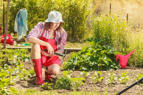 A woman dressed in a red apron, matching rain boots, and a wide-brimmed hat is kneeling in a well-maintained garden in Mayfair, tending to a variety of young plants and vegetable seedlings in the soil. The garden features neatly arranged beds with healthy green foliage, bordered by soil borders, and maintained grass pathways. In the background, there are flowering plants and shrubs, as well as a clothesline with hanging laundry, indicating a functional outdoor space. The scene is illuminated by natural sunlight, suggesting a bright, clear day, and the garden appears to be part of a landscaped outdoor area in an urban setting. The woman’s activity, along with the presence of garden tools and watering cans, reflects professional gardening and outdoor maintenance services offered by Landscapers Mayfair, focused on enhancing garden aesthetics and plant health in the local area. The overall setting encompasses a well-organized, environmentally conscious garden environment with a variety of textures and natural colour tones, suitable for garden renovation and landscaping projects within the London Mayfair postcode area.