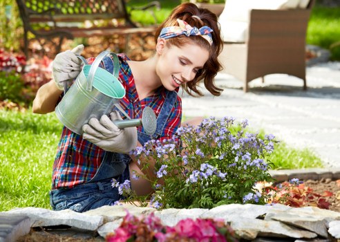 A young woman with light brown hair tied back with a colorful headband, wearing a blue and red checked shirt and grey gardening gloves, is kneeling in a well-maintained garden in Mayfair. She is watering a cluster of small, purple flowering plants within a curved stone edging boundary, with a joyful expression on her face. The garden features a lush green lawn with neatly trimmed grass, surrounded by flower beds containing various plants and shrubs. In the background, there is a paved patio area with outdoor furniture, including a wooden deck and a table, suggesting a landscaped outdoor space designed for relaxation and garden enjoyment. The scene indicates active gardening and plant care, aligning with services offered by Landscapers Mayfair, with a focus on well-organized planting schemes and outdoor aesthetic enhancement, set in a distinguished residential area that emphasizes natural beauty and landscape maintenance. The lighting suggests a bright, sunny day, highlighting natural tones and textures of the garden elements.