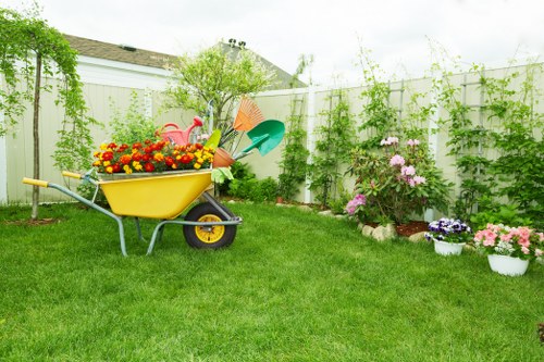 A neatly maintained residential garden featuring a vibrant green lawn with evenly cut grass, bordered by flower beds and potted plants. In the foreground, there is a yellow wheelbarrow filled with colourful garden flowers and gardening tools, positioned on the grassy area. To the right, flowering shrubs with pink and purple blossoms are planted alongside the white fence, which is decorated with climbing green vines. The background shows a small tree and a partly cloudy sky, suggesting a mild weather condition. The garden area is well-organized, with clear delineation between the lawn, flower beds, and potted plants, indicating ongoing or recent gardening maintenance that Landscapers Mayfair could provide as part of their services in the Mayfair district and postcode areas near central London. This outdoor space reflects a typical UK residential garden designed for aesthetic appeal and outdoor enjoyment, with attention to planting and tidy surfaces.