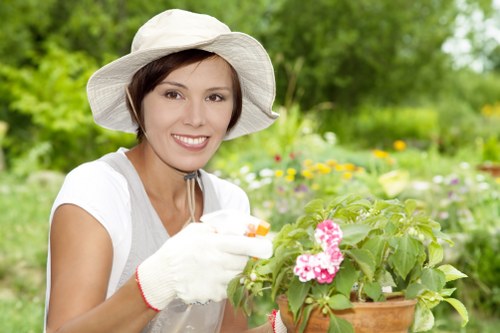 A woman in a white sun hat, light-colored gardening gloves, and a casual white top is smiling while tending to a potted flowering plant outdoors in a lush garden. The garden features a well-maintained lawn with dense green grass, bordered by a variety of flowering plants and shrubs. In the background, there are tall trees and additional planting areas, indicating a landscaped backyard or front garden space. The scene suggests a bright, sunny day with natural light illuminating the garden, and the woman appears to be engaged in gardening or planting activity, supporting outdoor maintenance services offered by Landscapers Mayfair in the local area.