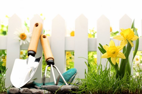 The image showcases a small section of a garden, featuring a white picket fence in the background with soft sunlight filtering through. In the foreground, there is a patch of vibrant green grass and a few blooming yellow daffodils on the right side, adding natural colour contrast. Near the fence, garden tools including a trowel and a transplanter with wooden handles and metal blades are resting on the soil, indicating active gardening. The soil appears healthy and slightly raised, bordered by the lush grass and flowering plants. The environment suggests a well-maintained outdoor space suitable for a variety of planting and landscaping activities, with natural light emphasizing the freshness of the greenery. This scene reflects typical garden care tasks such as planting, soil tending, and general outdoor maintenance that companies like Landscapers Mayfair offer within residential gardens in the Mayfair area, inferring local context from the scene and the service focus.