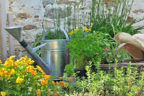 A vibrant garden scene featuring a variety of plants and flowers, including bright yellow and orange blooms in the foreground, with lush green foliage and herb plants behind them. A galvanized metal watering can is placed among the plants, and a straw hat rests on a wooden surface to the right, suggesting outdoor gardening activity. The background reveals a stone wall with climbing greenery, adding a rustic and natural aesthetic to the space. The overall composition showcases well-maintained flower beds and garden containers, typical of outdoor gardening efforts in the Mayfair area, with natural lighting indicating a clear, sunny day. This scene emphasizes the importance of garden maintenance and plant care, aligning with landscaping and gardening services provided by companies like Landscapers Mayfair, especially in residential outdoor spaces or backyard gardens near W1 postcode areas.