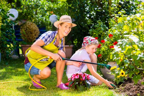 A woman and a young girl are gardening together in a well-maintained backyard garden, which features a lush, green lawn bordered by a variety of vibrant flowering plants and shrubs. The woman, wearing a straw hat, blue checkered shirt, and a bright yellow gardening apron, is smiling as she holds a garden hose, watering the flower beds. The girl, dressed in a pink top, light-colored pants, and a floral-patterned sun hat, is crouched down, carefully tending to the flowers with her hand. The garden includes a mix of colorful blooms such as yellow and red flowers, set against a background of leafy trees and bushes that provide shade and privacy. Decorative outdoor lighting elements are visible among the foliage, enhancing the garden's welcoming atmosphere. The scene suggests active outdoor maintenance and gardening care, typical of services offered by companies like landscapersmayfair.co.uk, emphasizing natural landscape elements, varied textures, and a vibrant, healthy outdoor environment in the Mayfair area, postcode W1 or nearby.