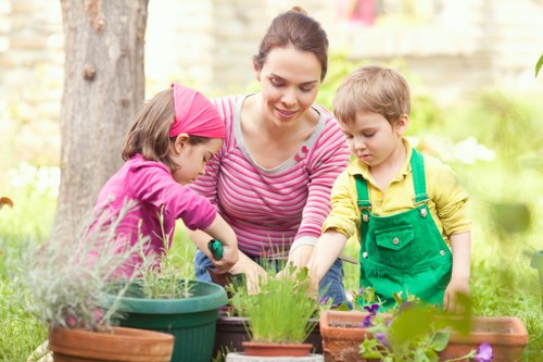 A woman with dark hair tied back, dressed in a pink and white striped long-sleeve top, is working alongside two children in a garden during daytime. The girl, wearing a pink headscarf and pink top, and the boy, dressed in a yellow shirt with green overalls, are engaged in planting or tending to potted plants on a wooden garden table. The garden features a lush, green lawn, with a mature tree trunk visible on the left side, and a background of a garden fence and low-growing shrubs. Various plant pots filled with soil and healthy green foliage are arranged on the table, which includes flowering plants and herbs, contributing pleasant natural colour contrasts. The scene is illuminated by soft, natural sunlight, suggesting a clear or slightly cloudy sky, creating a warm outdoor atmosphere suitable for gardening activities. This setting reflects a well-maintained outdoor space, typical of residential gardens in Mayfair, where landscaping services by Landscapers Mayfair might be employed to enhance garden structures and plant care, supporting a family-friendly environment.