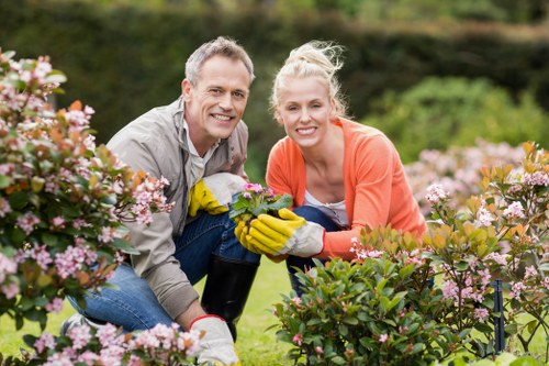 A middle-aged man and a young woman are kneeling together in a well-maintained garden, smiling at the camera while tending to vibrant pink and purple flowering bushes. The man is wearing a light grey shirt and yellow gardening gloves, holding a small gardening tool, while the woman is dressed in a coral long-sleeved top and also wears yellow gloves. Behind them, the garden features a lush green lawn, a backdrop of trees, and a neatly trimmed hedge line, all under natural daylight with a clear sky. The scene showcases outdoor gardening activities, highlighting the care of shrubbery and flowerbeds, characteristic of professional gardening and landscaping services in Mayfair. Their actions demonstrate pruning and maintenance, contributing to an attractive, well-organized outdoor space suitable for garden design and upkeep in the local area.
